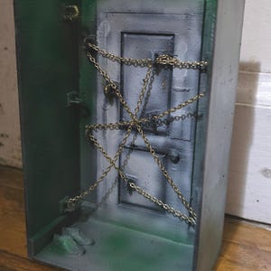 Puede incluir: Un diorama en miniatura de una puerta asegurada con cadenas. La escena está pintada en tonos de verde y gris, con una cadena dorada que cruza la puerta. El efecto general es un tema de casa embrujada espeluznante.