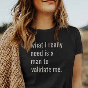 May include: A black t-shirt with the text "what I really need is a man to validate me." The model is wearing the shirt with a brown cardigan and a black belt.
