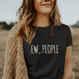 May include: A woman wearing a black t-shirt with the text "EW, PEOPLE" in white lettering. She is wearing a brown cardigan and a black belt with a silver buckle. The woman is standing in a field of tall grass.