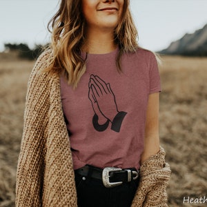 Puede incluir: Camiseta de color malva jaspeado con un gr&aacute;fico negro de manos rezando. La camiseta de cuello redondo se usa con una rebeca beige y pantalones negros con una hebilla plateada. El fondo es un campo. El texto "Heather Mauve" es visible.