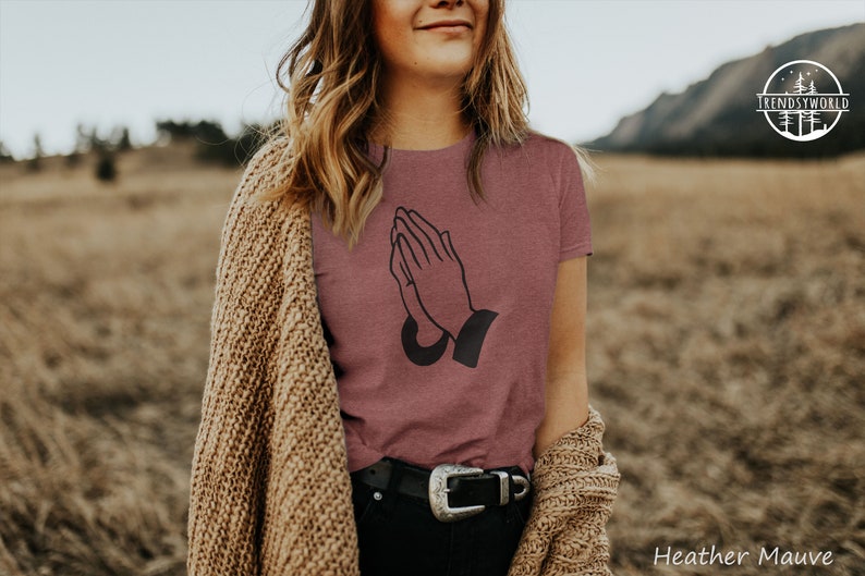 Puede incluir: Camiseta de color malva jaspeado con un gr&aacute;fico negro de manos rezando. La camiseta de cuello redondo se usa con una rebeca beige y pantalones negros con una hebilla plateada. El fondo es un campo. El texto "Heather Mauve" es visible.