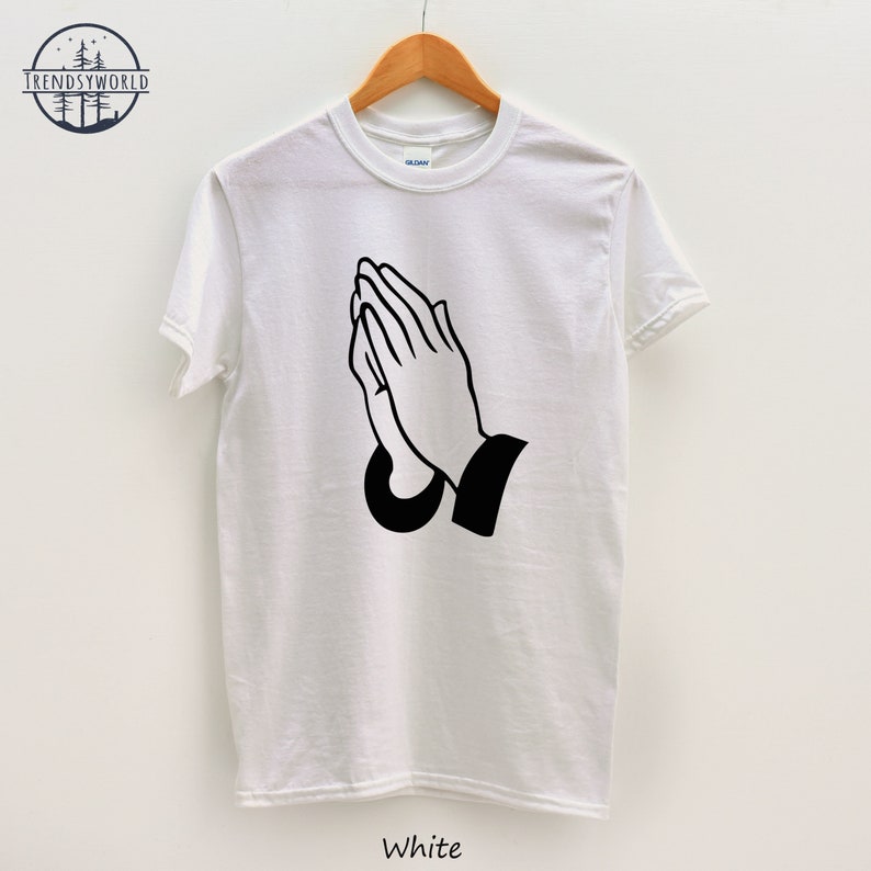 Puede incluir: Camiseta blanca con un gr&aacute;fico negro de manos rezando. La camiseta est&aacute; colgada en una percha de madera. La palabra "White" est&aacute; impresa en la parte inferior de la camiseta.