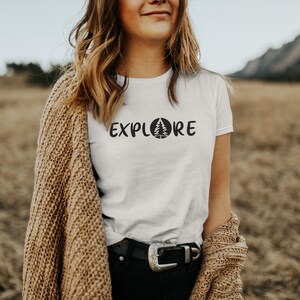 Pu&ograve; includere: Maglietta bianca con il testo "EXPLORE" e una grafica di pino stilizzata. La modella indossa un cardigan marrone e si trova in un campo.