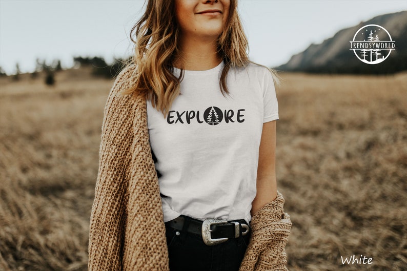 Pu&ograve; includere: Maglietta bianca con il testo "EXPLORE" e una grafica di pino stilizzata. La modella indossa un cardigan marrone e si trova in un campo.