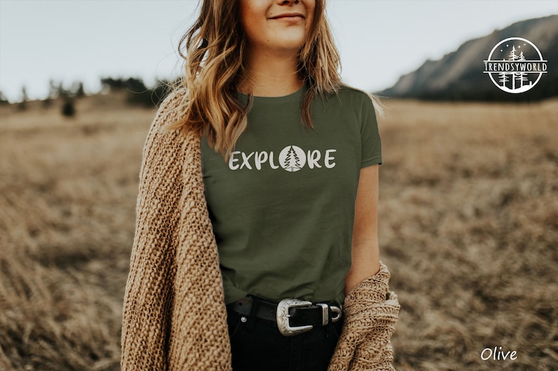 Pu&ograve; includere: Una donna indossa una maglietta verde con la scritta "EXPLORE" e un'immagine di un pino. Indossa un cardigan marrone e una cintura nera con una fibbia argentata. Lo sfondo &egrave; un campo di erba alta.