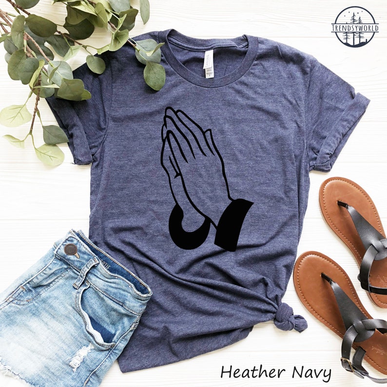 Puede incluir: Camiseta azul marino jaspeada con un gr&aacute;fico negro de manos rezando. La imagen incluye pantalones cortos de mezclilla y sandalias marrones. El texto "Heather Navy" est&aacute; impreso en la parte inferior.