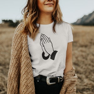 Puede incluir: Camiseta blanca con un gr&aacute;fico negro de manos rezando. El atuendo incluye pantalones negros y un c&aacute;rdigan marr&oacute;n. El texto "TRENDSYWORLD" y "White" est&aacute;n incluidos.