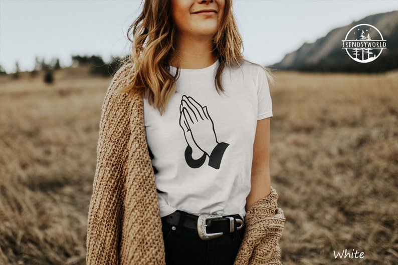 Puede incluir: Camiseta blanca con un gr&aacute;fico negro de manos rezando. El atuendo incluye pantalones negros y un c&aacute;rdigan marr&oacute;n. El texto "TRENDSYWORLD" y "White" est&aacute;n incluidos.