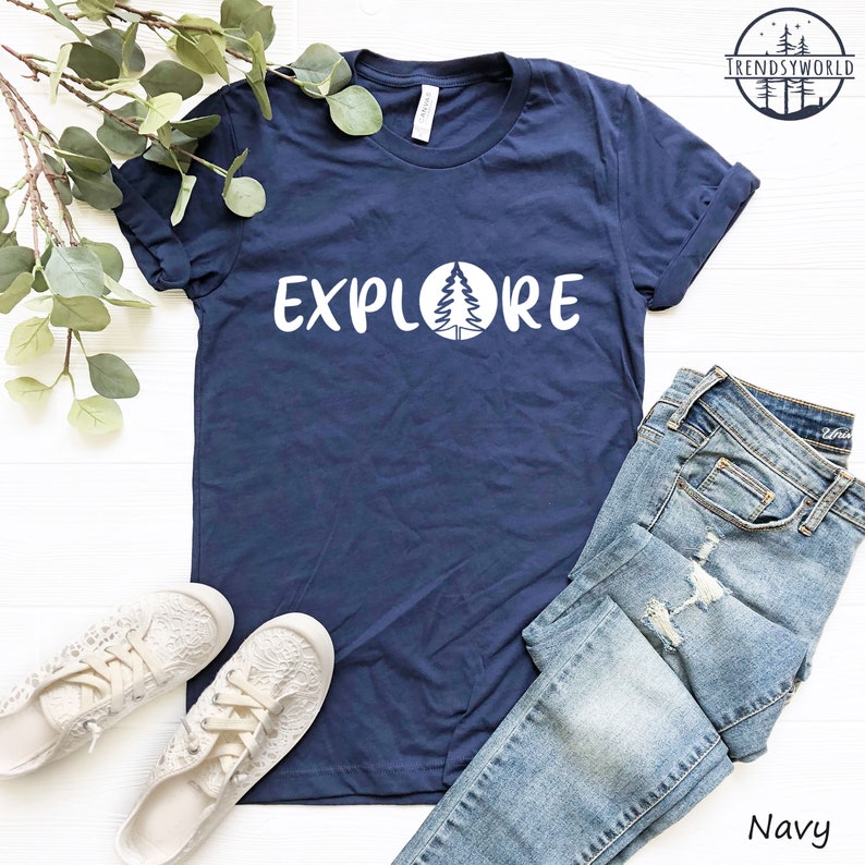 Pu&ograve; includere: Una maglietta blu navy con la parola "EXPLORE" stampata in bianco con un pino stilizzato all'interno di un cerchio. La maglietta &egrave; stesa su una superficie bianca con un paio di scarpe di tela bianche e un paio di jeans blu.