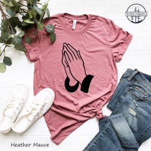Puede incluir: Camiseta color malva jaspeado con un gr&aacute;fico negro de manos rezando. La camiseta se combina con vaqueros azules y zapatillas blancas con cordones. El texto "Heather Mauve" est&aacute; impreso en la parte inferior de la imagen.