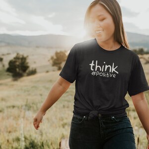 Puede incluir: Una persona lleva una camiseta gris oscuro con el texto "think #positive" en letras blancas. La persona camina por un campo con hierba alta y una cordillera de monta&ntilde;as al fondo.