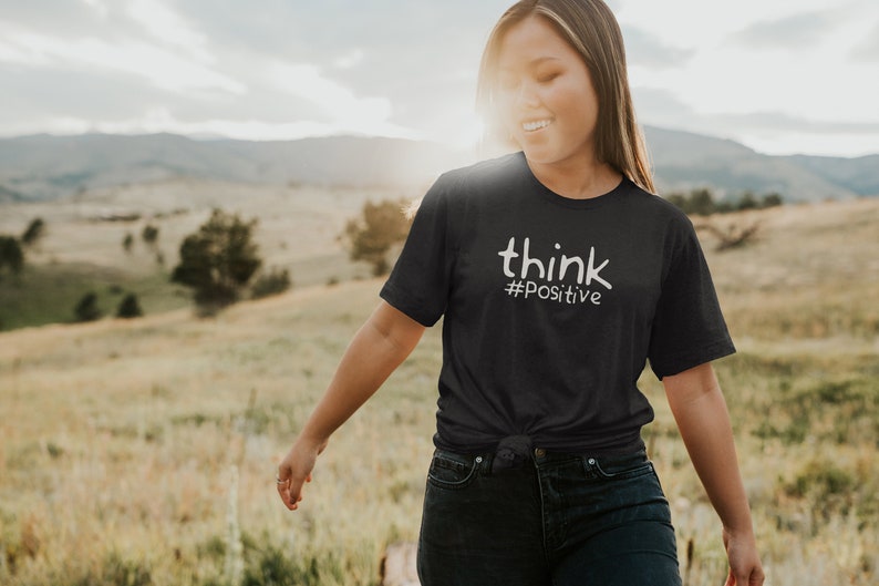 Puede incluir: Una persona lleva una camiseta gris oscuro con el texto "think #positive" en letras blancas. La persona camina por un campo con hierba alta y una cordillera de monta&ntilde;as al fondo.