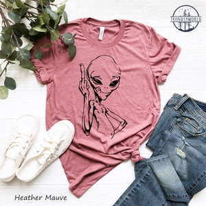 K&ouml;nnte beinhalten: Heather-mauvefarbenes T-Shirt mit einem schwarzen Aufdruck eines Aliens, das den Mittelfinger zeigt. Das Shirt liegt flach auf einer wei&szlig;en Oberfl&auml;che mit einem Paar wei&szlig;en Schuhen und einer blauen Jeans.