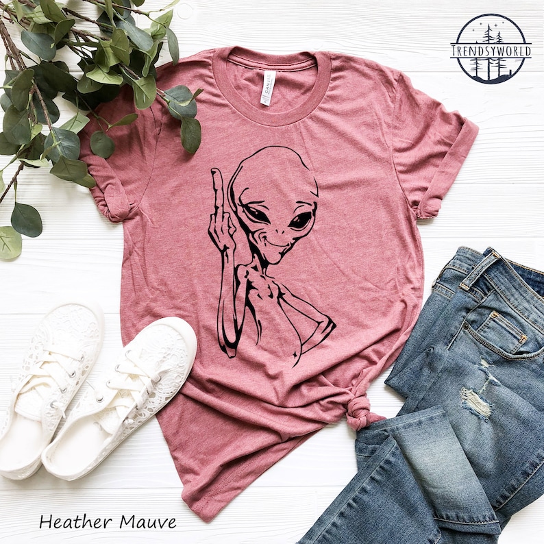 K&ouml;nnte beinhalten: Heather-mauvefarbenes T-Shirt mit einem schwarzen Aufdruck eines Aliens, das den Mittelfinger zeigt. Das Shirt liegt flach auf einer wei&szlig;en Oberfl&auml;che mit einem Paar wei&szlig;en Schuhen und einer blauen Jeans.