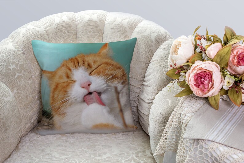 May include: A close-up of a ginger cat with white markings licking its paw. The cat is lying on a light blue pillow with its eyes closed. The pillow is on a white armchair.