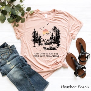 Puede incluir: Una camiseta de color melocotón con un gráfico de una camioneta vintage conduciendo por un bosque con una montaña y una puesta de sol en el fondo. El texto "Let's just go and not come back for a while" está impreso debajo del gráfico.