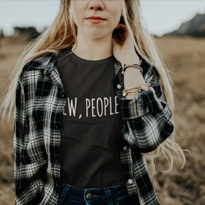 May include: A woman wearing a black t-shirt with the text "Few, People" in white lettering. She is wearing a plaid shirt over the t-shirt and has long blonde hair.