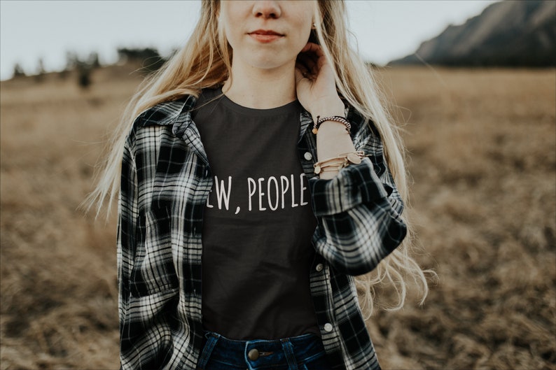 May include: A woman wearing a black t-shirt with the text "Few, People" in white lettering. She is wearing a plaid shirt over the t-shirt and has long blonde hair.