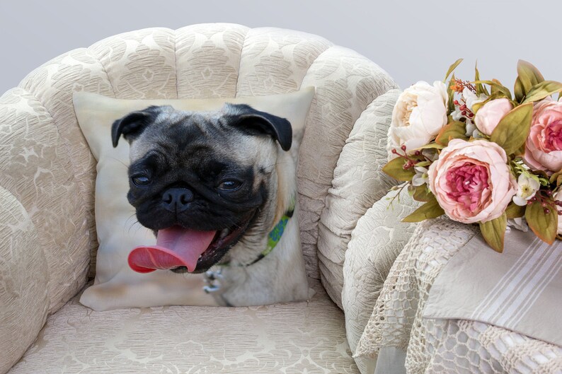 May include: A tan and black pug dog with a pink tongue sticking out is printed on a white pillow. The pillow is on a white armchair with a floral bouquet on a table next to it.