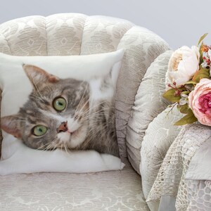 May include: A gray and white cat with green eyes is lying on a white pillow. The pillow is on a white armchair with a floral pattern. A bouquet of pink and white flowers is on the arm of the chair.