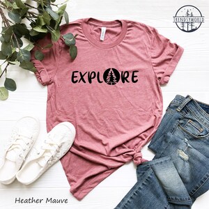 Pu&ograve; includere: Maglietta color malva heather con la parola "EXPLORE" stampata in nero con un albero all'interno di un cerchio. La maglietta &egrave; stesa su una superficie bianca con un paio di scarpe bianche e un paio di jeans blu.