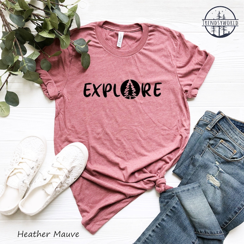 Pu&ograve; includere: Maglietta color malva heather con la parola "EXPLORE" stampata in nero con un albero all'interno di un cerchio. La maglietta &egrave; stesa su una superficie bianca con un paio di scarpe bianche e un paio di jeans blu.