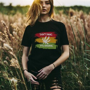May include: A woman wearing a black t-shirt with a green, yellow, and red graphic that says "Don't Panic It's Organic" and a white marijuana leaf. She is standing in a field of tall grass.