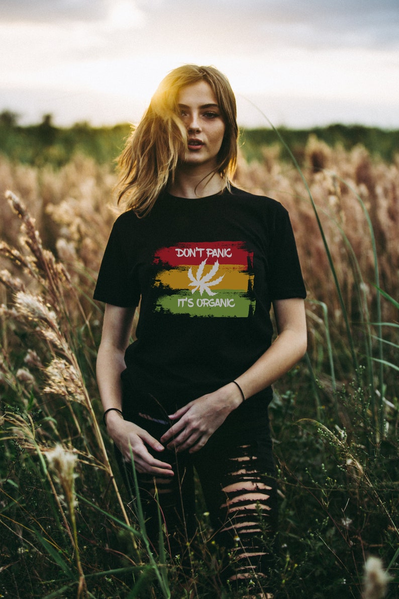 May include: A woman wearing a black t-shirt with a green, yellow, and red graphic that says "Don't Panic It's Organic" and a white marijuana leaf. She is standing in a field of tall grass.