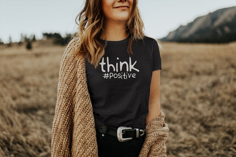 Puede incluir: Una camiseta negra con el texto "think #positive" en letras blancas.