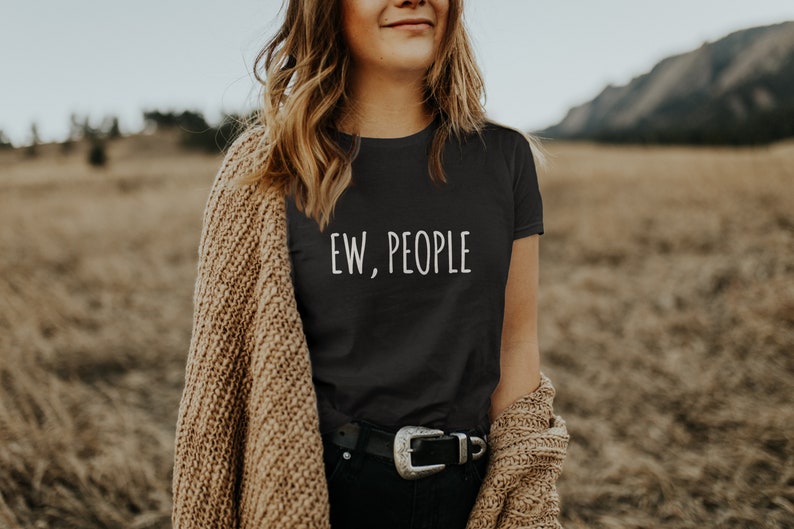 May include: A woman wearing a black t-shirt with the text "EW, PEOPLE" in white lettering. She is wearing a brown cardigan and a black belt with a silver buckle. The woman is standing in a field of tall grass.