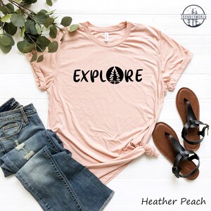 Pu&ograve; includere: Una maglietta color pesca con il testo "EXPLORE" in nero con una silhouette nera di un pino all'interno di un cerchio. La maglietta &egrave; stesa su una superficie bianca con un paio di sandali marroni e un paio di jeans blu.