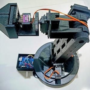 Può includere: Un braccio robotico nero con più giunti e una base circolare. Il braccio ha cablaggio arancione e una piccola scheda di circuito. Il braccio è realizzato in plastica nera ed è progettato per una varietà di compiti.