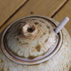 Könnte beinhalten: Eine Keramik-Zuckerdose mit Deckel und kleinem Löffel. Die runde Schale hat ein strukturiertes, wabenartiges Muster in Beige, Braun und Orange. Der Deckel hat einen sechseckigen Knopf. Der Löffel ist hellgrau.