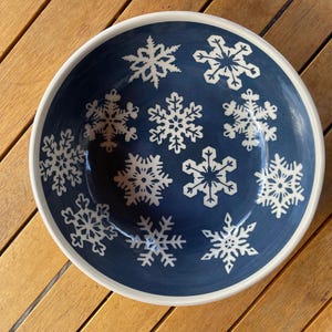 Puede incluir: Un cuenco redondo de cerámica con interior azul marino y borde blanco. El cuenco está decorado con diseños de copos de nieve blancos. El cuenco está sobre una superficie de madera.