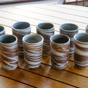 Könnte beinhalten: Acht hohe Keramikbecher mit einem braunen und weißen Wirbelmuster. Das Innere jedes Bechers ist in einem gedämpften Grauton gehalten. Die Becher sind auf einem Holztisch angeordnet.