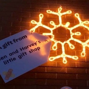 May include: A white sign with the text "A gift from Ben and Harvey's little gift shop" is attached to a brick wall with a glowing orange snowflake behind it.