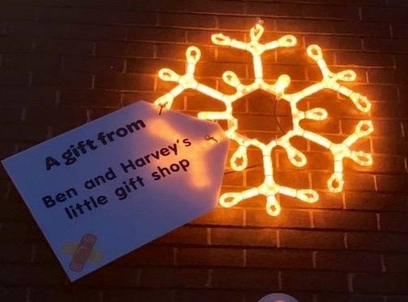 May include: A white sign with the text "A gift from Ben and Harvey's little gift shop" is attached to a brick wall with a glowing orange snowflake behind it.