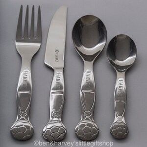May include: A set of four silver-toned children's cutlery with a soccer ball design on the handles. The knife is engraved with "VINERS" and the fork, spoon, and teaspoon are engraved with "HARVEY".