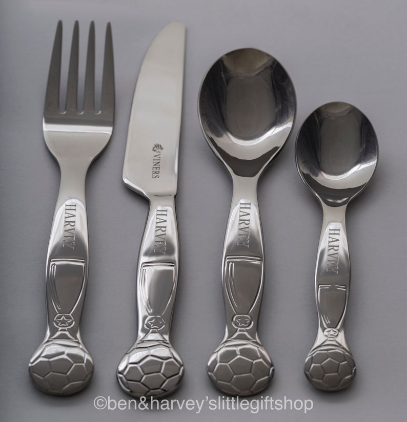 May include: A set of four silver-toned children's cutlery with a soccer ball design on the handles. The knife is engraved with "VINERS" and the fork, spoon, and teaspoon are engraved with "HARVEY".