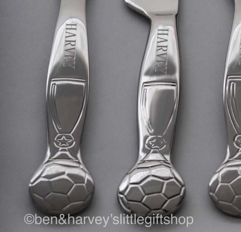 May include: Three silver-colored, personalized children's cutlery sets with a soccer ball design. Each handle is engraved with the name "Harvey".