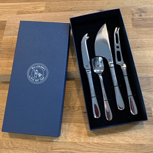 Może przedstawiać: Zestaw czterech srebrnych noży do sera w niebieskim pudełku z aksamitu. Noże są oznaczone napisem "The Cheese Knife Shop" i przedstawiają małą ilustrację osoby i psa.