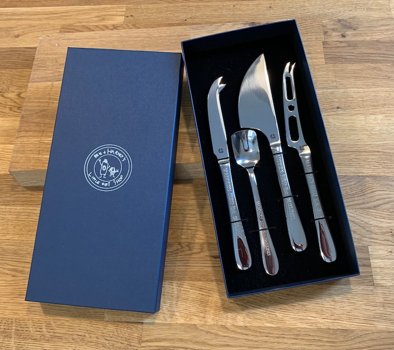 Może przedstawiać: Zestaw czterech srebrnych noży do sera w niebieskim pudełku z aksamitu. Noże są oznaczone napisem "The Cheese Knife Shop" i przedstawiają małą ilustrację osoby i psa.