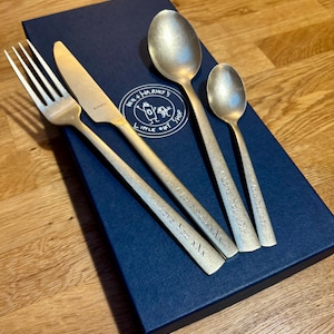 May include: A set of four brushed gold-colored utensils: a fork, knife, and two spoons. The utensils are engraved with "I Love You XXX" on the handles. The set rests on a navy blue box with a circular logo.