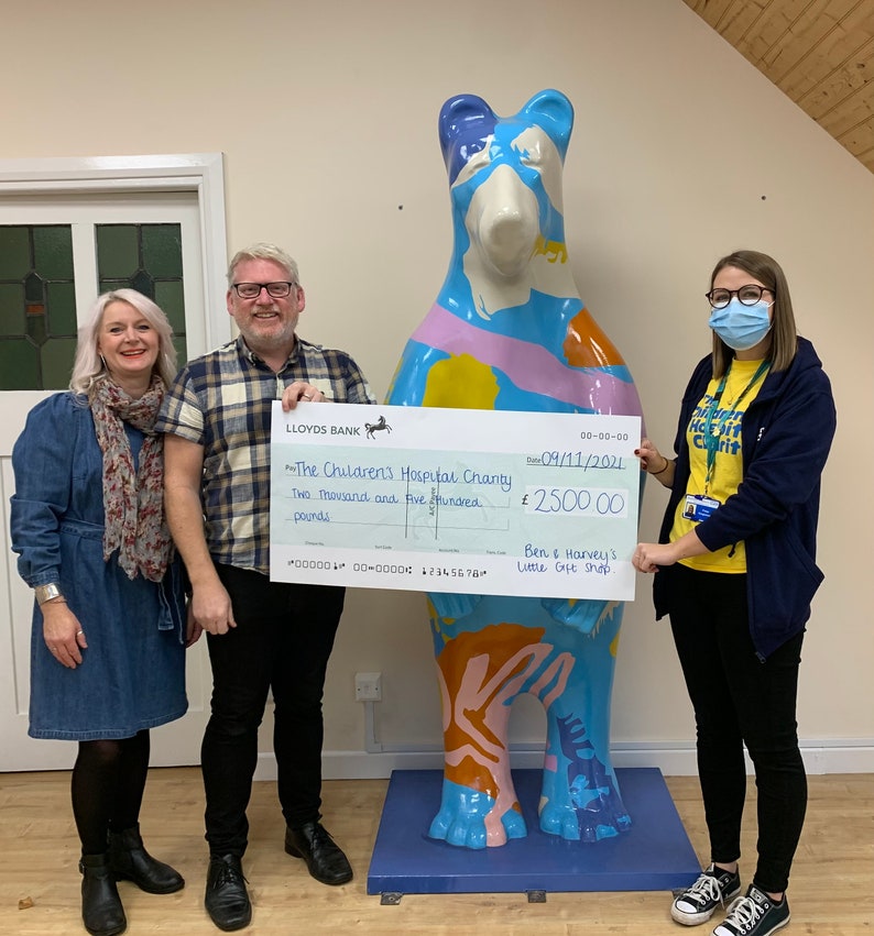 May include: A check for two thousand five hundred pounds is being presented to The Children's Hospital Charity by Ben & Harvey's Little Gift Shop. The check is dated 09/11/2021. The check is being presented in front of a colorful bear sculpture.