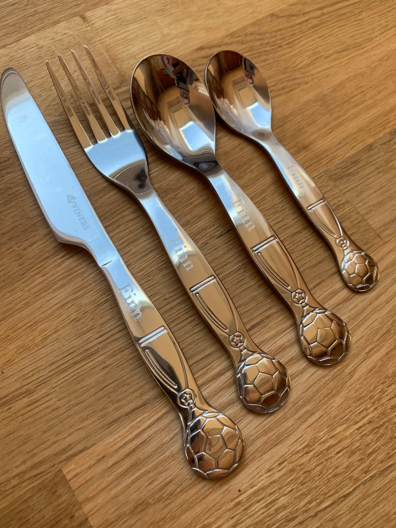 May include: Set of four silver-toned utensils with a soccer ball design on the handles. The utensils include a knife, fork, and two spoons. The words "Kim" and "Winners" are engraved on the handles.