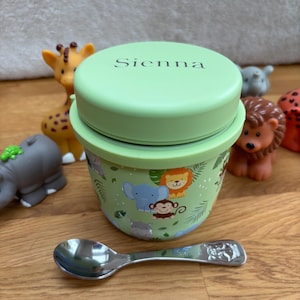 May include: A light green baby food container with a matching lid, featuring the name "Sienna". The container is decorated with cartoon animals. A silver spoon rests in front of the container. Toy animals are in the background.