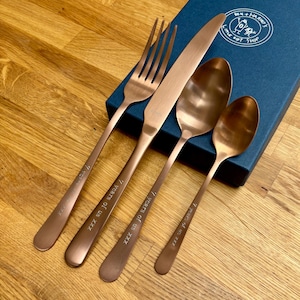 May include: A copper-colored flatware set including a fork, knife, and two spoons. The handles are engraved with "7 years of us XXX". The set is next to a blue gift box with the text "BEN + HARRI'S LITTLE GIFT SHOP".