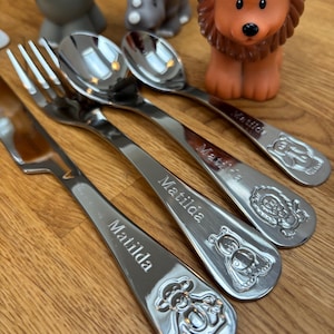 May include: A set of four stainless steel baby utensils, including a fork and three spoons, each engraved with the name "Matilda" and a different animal design. Toy animal figures are in the background.