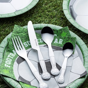 May include: A set of silver football-themed cutlery on a green and white paper plate with a soccer ball design. The cutlery includes a fork, knife, and two spoons.