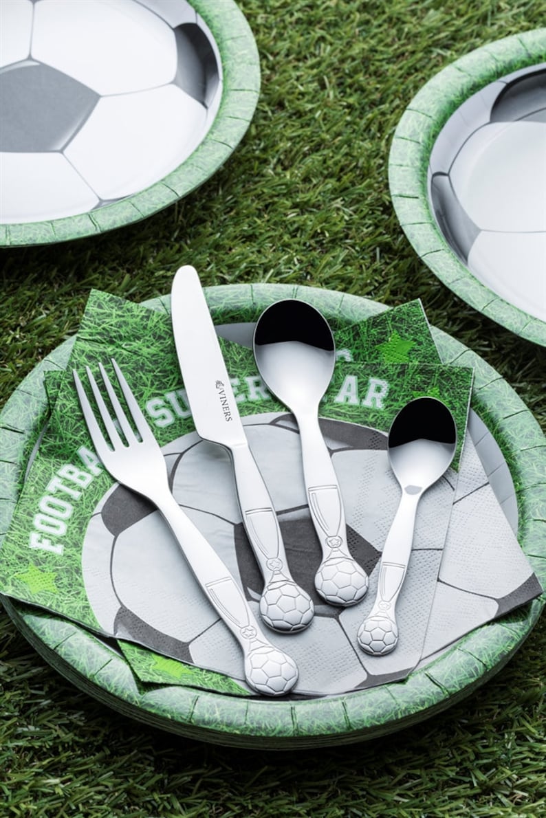 May include: A set of silver football-themed cutlery on a green and white paper plate with a soccer ball design. The cutlery includes a fork, knife, and two spoons.